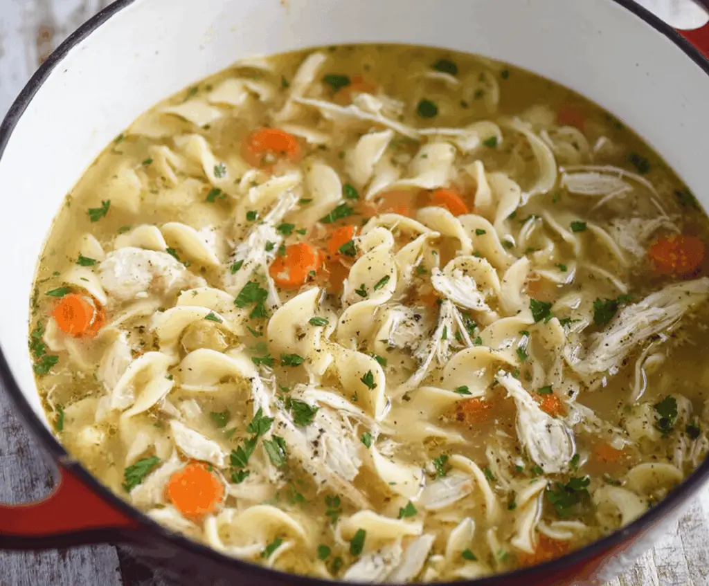 A steaming bowl of CopyCat Panera Bread Chicken Noodle Soup with tender chicken, carrots, celery, and egg noodles garnished with fresh herbs, served in a white bowl on a wooden table.
