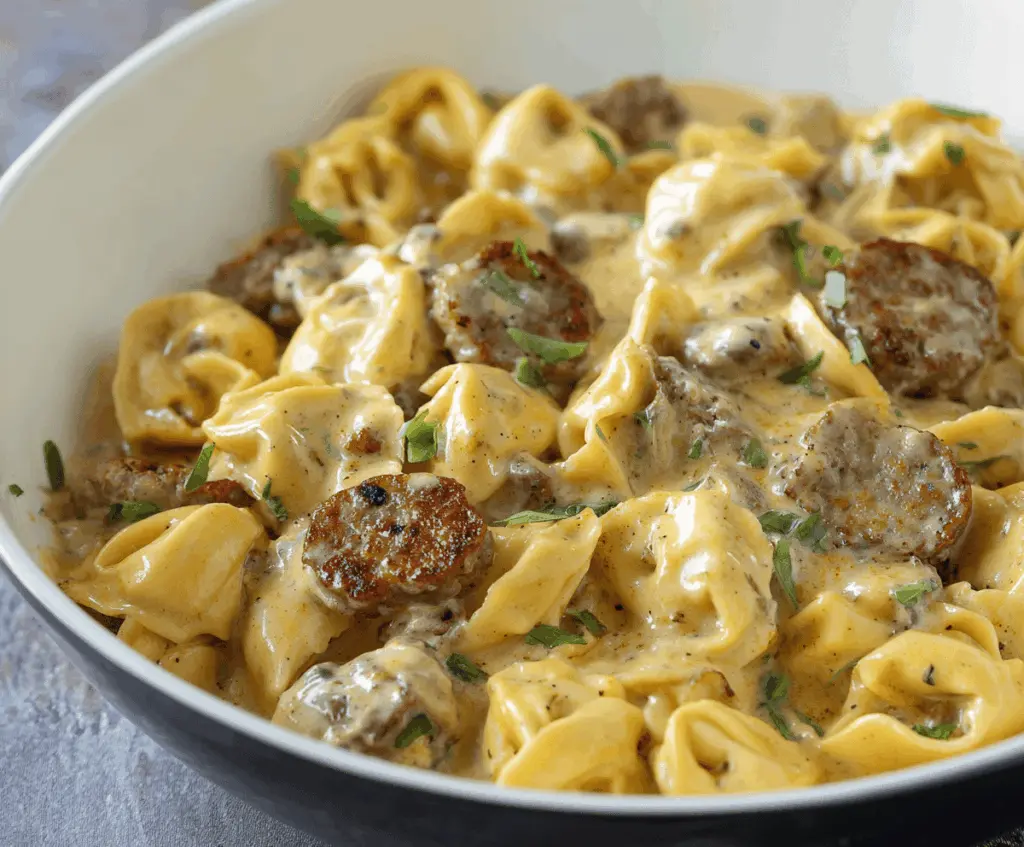 Creamy sausage and cheese tortellini Alfredo dish with grilled sausage slices, topped with fresh herbs and Parmesan cheese, served in a white bowl.